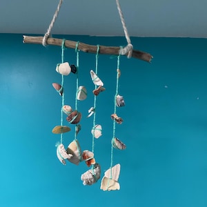 May include: A handmade seashell mobile. A natural wooden branch suspends various seashells of different shapes and colors, strung on teal-colored twine. The mobile hangs from a white rope against a blue background.