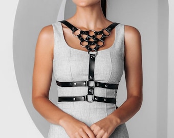 Puede incluir: Un arnés de cuero negro con anillos y hebillas de metal. El arnés se lleva sobre un vestido gris sin mangas.