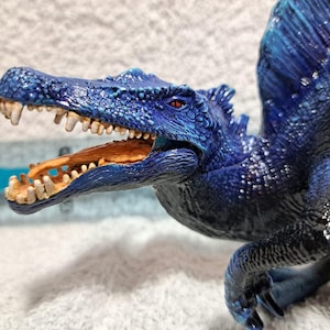 Könnte beinhalten: Detaillierte Figur eines Spinosaurus-Dinosauriers, überwiegend blau mit dunkleren Schattierungen. Der Dinosaurier hat ein strukturiertes, geschupptes Aussehen mit einem markanten Segel auf dem Rücken. Sein Maul ist geöffnet und zeigt Zähne und ein braunes Inneres.