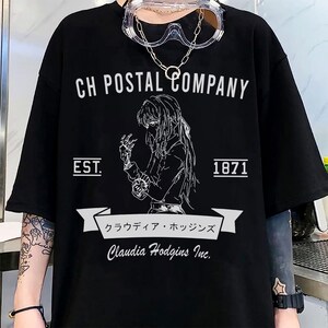 May include: Black t-shirt with a white graphic featuring the text "CH POSTAL COMPANY" above an illustration of a person. Below the illustration is the text "Claudia Hodgins Inc." and Japanese text. The shirt also has "EST. 1871".