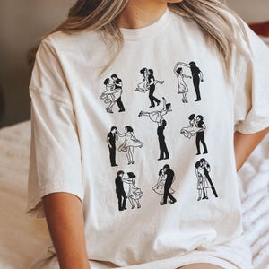 Puede incluir: Una camiseta blanca con un estampado gr&aacute;fico negro de una pareja bailando en varias poses. El gr&aacute;fico se repite nueve veces en la camiseta.