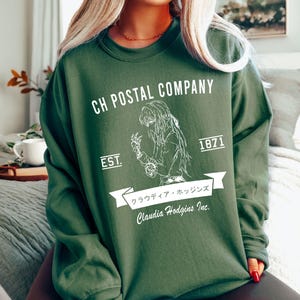 May include: Dark green sweatshirt with white graphic text and illustration. The text reads "CH POSTAL COMPANY" above an illustration of a person. Below the illustration is the text "Claudia Hodgins Inc." and Japanese text.