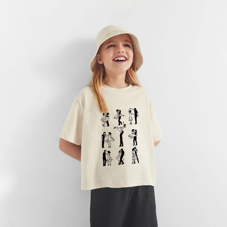Puede incluir: Una persona joven lleva una camiseta de color crema con un estampado gr&aacute;fico en blanco y negro de parejas bailando. La camiseta tiene mangas cortas y cuello redondo. La persona tambi&eacute;n lleva un sombrero de cubo negro y pantalones negros.
