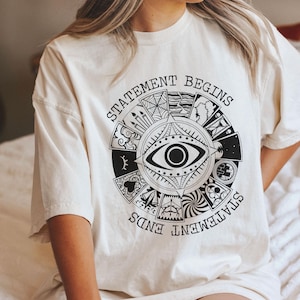 May include: A white t-shirt with a black and white graphic print of an eye surrounded by a circle of symbols. The text "Statement Begins" and "Statement Ends" is printed around the circle.