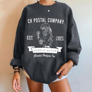 May include: Dark gray sweatshirt with white graphic text and illustration. The text reads "CH POSTAL COMPANY" above an illustration of a person. Below the illustration is a banner with Japanese text and the name "Claudia Hodgins Inc." The sweatshirt also has the text "EST. 1871".