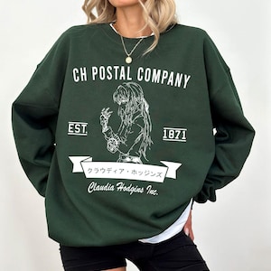 May include: Dark green sweatshirt with white text and graphic. The text reads "CH POSTAL COMPANY" above a detailed illustration of a person. Below the illustration is a banner with Japanese text and "Claudia Hodgins Inc." The sweatshirt also has "EST. 1871".