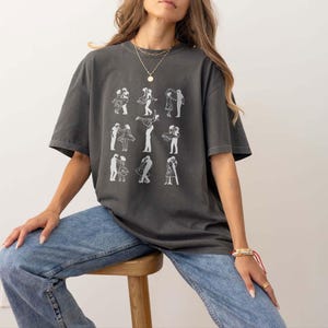 Puede incluir: Una camiseta gris de gran tama&ntilde;o con un gr&aacute;fico blanco de parejas bailando. La camiseta la lleva puesta una persona con el pelo largo y casta&ntilde;o y se combina con unos vaqueros azules.