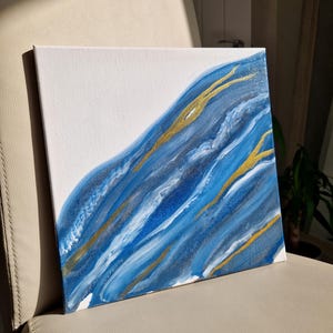 Könnte beinhalten: Abstraktes Gemälde auf Leinwand mit blauer, weißer und goldener Farbe. Die Farbe ist verwirbelt und geschichtet, um einen dynamischen und strukturierten Effekt zu erzielen.
