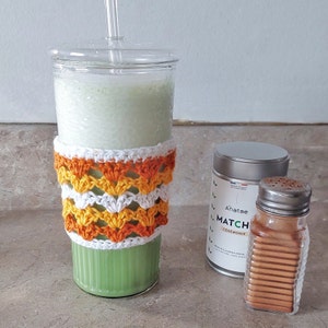 Peut inclure: Un smoothie vert dans un verre transparent avec un manchon de tasse en crochet blanc et orange. Le smoothie est surmonté d'une paille transparente. Un récipient en argent de thé matcha et un bocal en verre d'épices brunes sont sur le comptoir.