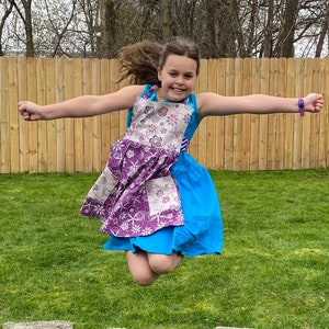 May include: A young person wearing a purple and white floral print dress with a blue overlay and a purple bracelet. The person is jumping in the air with their arms outstretched.