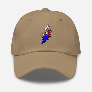 May include: A tan baseball cap featuring a colorful graphic of a skeleton wearing a top hat, riding a red, white, and blue lightning bolt. The cap has a curved brim and a button on top.