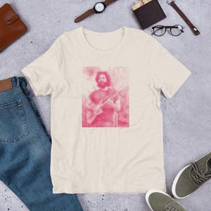 May include: A light beige t-shirt with a pink pixelated image of a person playing a guitar.