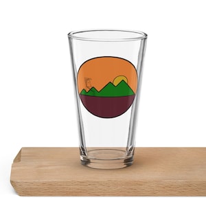 May include: A clear glass pint glass with a round sticker featuring a sunset over green mountains and a tower in the foreground. The sticker has a maroon border.
