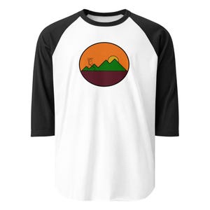 Könnte beinhalten: Ein weißes Baseball-Shirt mit schwarzen Ärmeln und einer kreisförmigen Grafik eines Sonnenuntergangs über grünen Bergen. Die Grafik hat einen kastanienbraunen Hintergrund und eine orange Sonne mit gelbem Zentrum.