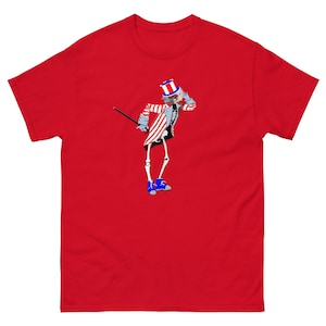May include: Red t-shirt featuring a graphic of a skeleton dressed as Uncle Sam. The skeleton is holding a cane and wearing a blue and white striped hat.