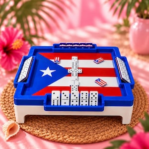 May include: A square domino game board featuring the Puerto Rican flag design in red, white, and blue. The blue plastic frame includes built-in racks for white domino tiles with black pips. Several dominoes are placed on the board surface.