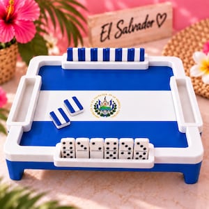 May include: A square domino game board featuring the blue and white flag of El Salvador with its central coat of arms. The board has white plastic racks on all four sides, with blue and white domino tiles placed in the racks and on the surface.