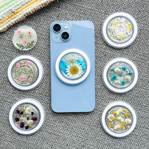 Puede incluir: Un teléfono inteligente azul claro con un agarre blanco adjunto, rodeado de seis agarres adicionales. Cada agarre presenta un diseño de resina transparente con flores prensadas en varios colores, incluyendo rosa, amarillo y azul. Los agarres tienen un borde blanco.