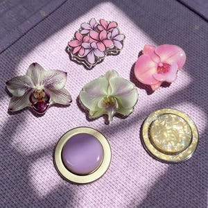Könnte beinhalten: Eine Sammlung von sechs dekorativen Handyhaltern mit verschiedenen Designs: ein Bündel aus rosa und violetten Blumen, eine rosa Orchidee, eine weiße Orchidee mit violetten Streifen, eine blassgrüne Orchidee, ein lavendelfarbener Kreis und ein cremefarbener Perlmuttkreis.