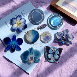Op de afbeelding: Een collectie blauwe telefoonhouders met diverse ontwerpen, waaronder geperste bloemen, een agaatplak, een vlinder en orchideeën. De items zijn gerangschikt op een lichtblauwe kaart met de tekst LOVE en SMILE EVERY DAY.