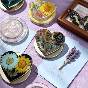 Peut inclure: Une collection de dessous de verre en résine en forme de cœur présentant divers motifs, notamment des tournesols jaunes pressés, du feuillage vert, des motifs floraux complexes en vert et or, et une combinaison de marguerite bleue et tournesol.