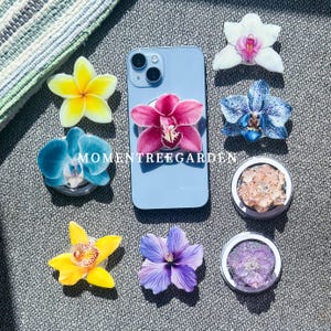 May include: A light blue smartphone with a pink orchid phone grip is surrounded by various flower-shaped phone grips in yellow, blue, purple, and white. Two circular grips contain dried flowers. The text "MOMENTREEGARDEN" is visible.