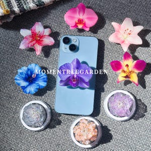 Może przedstawiać: Jasnoniebieski smartfon z przymocowanym fioletowym uchwytem na telefon w kształcie orchidei, otoczony różnymi uchwytami na telefon w kształcie kwiatów i prawdziwymi kwiatami. Uchwyty na telefon mają różne wzory kwiatowe, w tym różowe, niebieskie i żółte kwiaty. Widoczny jest tekst "MOMENTREEGARDEN".