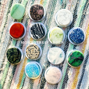 Könnte beinhalten: Eine Sammlung von zwölf runden Handyhaltern aus Stein in verschiedenen Farben und Mustern, darunter Grün, Rot, Schwarz und Weiß sowie Blau. Die Halterungen sind in silberfarbene Metallringe gefasst. Der Text "MOMENTREEGARDEN" ist sichtbar.