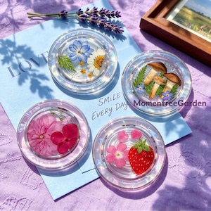 May include: Four circular clear resin coasters featuring embedded botanical elements. One contains blue and white flowers, another features three brown mushrooms, a third displays pink flowers, and the fourth includes a strawberry and pink petals.