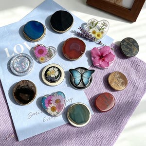 May include: A collection of circular and heart-shaped phone grips featuring various designs including polished agate slices in blue, red, black, and green, pressed flowers in resin, a blue butterfly graphic, and a pink hibiscus flower attachment.