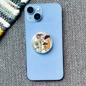 May include: A light blue smartphone with a clear, round phone grip attached. The grip features a design of dried mushrooms and small pink and white flowers encased in resin. The phone has a dual-lens camera.