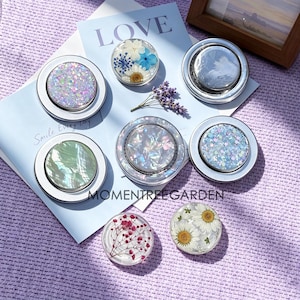 Peut inclure: Un ensemble de sept supports de téléphone circulaires présentant divers motifs, notamment des fleurs pressées, des effets de coquillage irisés et des paillettes multicolores. Les supports ont des bases argentées sur un papier bleu clair.