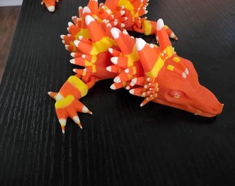 May include: A 3D-printed orange dragon figurine with a candy corn design. The dragon has a long, curved body, with white, yellow, and orange accents. The figurine is on a black surface.