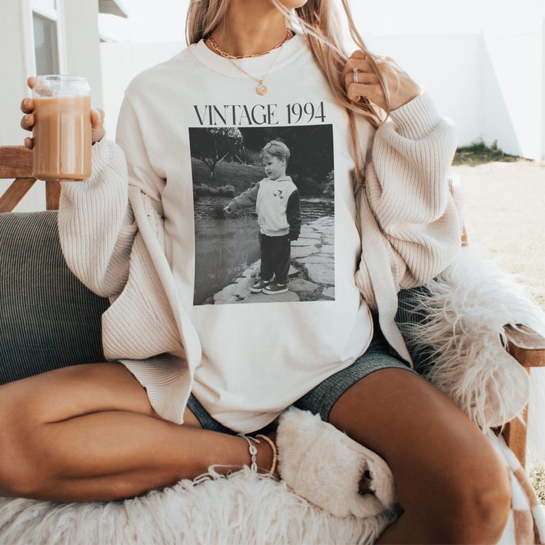 May include: Cream-colored sweatshirt with the text "VINTAGE 1994" above a black and white photo of a child. The sweatshirt is worn with shorts and a cream-colored cardigan. A glass of iced coffee is held in the hand.