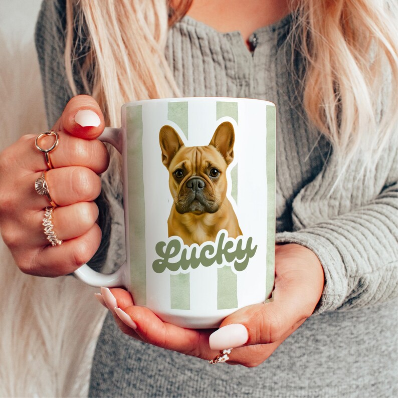 May include: White ceramic mug with a light green striped pattern and a brown French Bulldog graphic. The word "Lucky" is written in a green script font below the dog. The mug is held by a person.