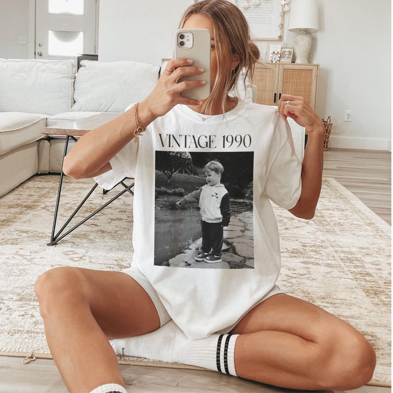 May include: White t-shirt with the text "VINTAGE 1990" above a black and white photo of a child. The child is wearing a hoodie and pants, standing near water. The shirt is being held up.