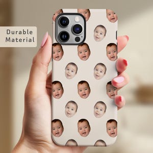 May include: A white phone case with a repeating pattern of baby faces. The phone case is designed for an iPhone 14 Pro Max. The case has a clear camera cutout and a black camera bump.