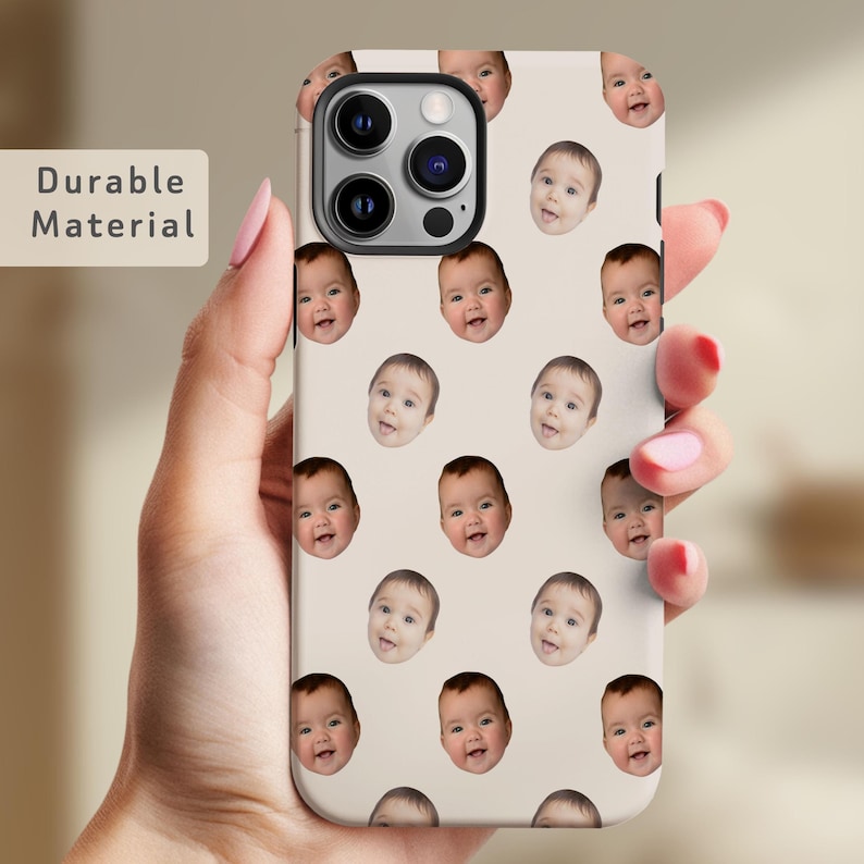 May include: A white phone case with a repeating pattern of baby faces. The phone case is designed for an iPhone 14 Pro Max. The case has a clear camera cutout and a black camera bump.
