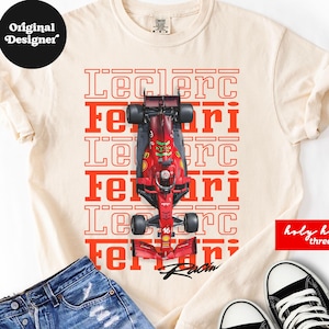 May include: A cream-coloured t-shirt featuring a top-down graphic of a red Formula 1 race car centred over repeating red text that reads Leclerc and Ferrari. The shirt is paired with blue denim shorts and black and white high-top trainers.