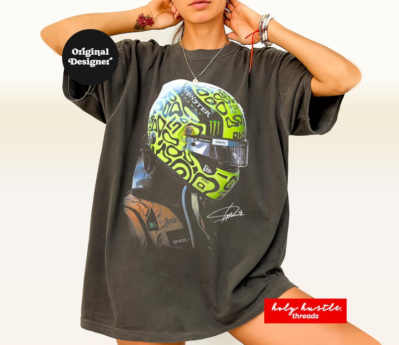 Puede incluir: Camiseta gris oscuro con un dise&ntilde;o de casco de carreras con detalles en verde ne&oacute;n y negro. La camiseta tiene un gr&aacute;fico de un piloto y los textos "Original Designer" y "holy hustle. threads."