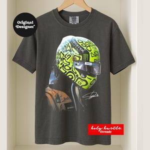 Puede incluir: Camiseta gris oscuro con un casco de carreras con un dise&ntilde;o verde ne&oacute;n y negro. El casco tiene el logotipo de Monster Energy. La camiseta tambi&eacute;n tiene el texto "Original Designer" y "holy hustle threads".