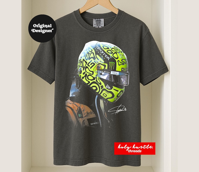 Puede incluir: Camiseta gris oscuro con un casco de carreras con un dise&ntilde;o verde ne&oacute;n y negro. El casco tiene el logotipo de Monster Energy. La camiseta tambi&eacute;n tiene el texto "Original Designer" y "holy hustle threads".