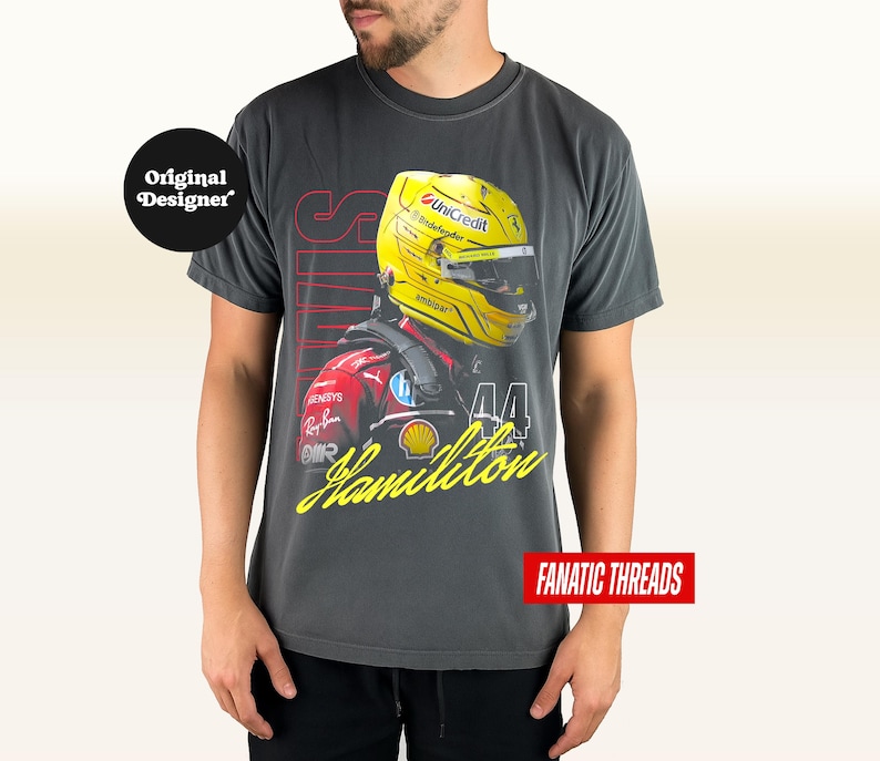 May include: Dark grey t-shirt with a graphic of a Formula 1 driver wearing a yellow helmet. The design includes the name "Hamilton" in yellow script, the number "44", and the word "SPEED" in red. The shirt has the text "FANATIC THREADS".