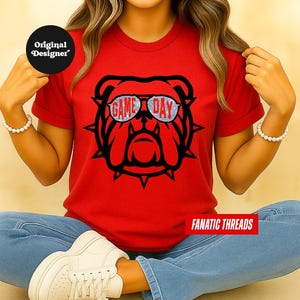 May include: A red t-shirt with a black bulldog graphic wearing sunglasses with the words "GAME DAY" in silver glitter. The shirt is from "FANATIC THREADS". A black circle with "Original Designer" is in the upper left.