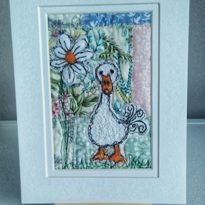 May include: A white framed artwork featuring a white embroidered goose with orange feet standing in a field of flowers. The flowers are embroidered on a patchwork of colorful fabrics.