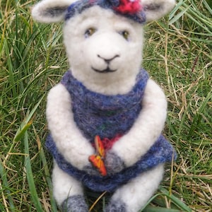 Puede incluir: Una muñeca de oveja de fieltro blanco que lleva un vestido azul con una diadema de flores rojas y sostiene una mariposa. La muñeca está sentada en un lecho de hierba verde.