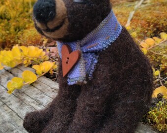 May include: A brown felt bear wearing a purple and blue plaid scarf with a brown heart-shaped button. The bear is sitting on a wooden surface.