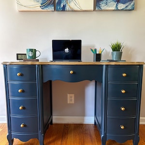 May include: A navy blue desk with gold hardware and a light wood top. The desk has multiple drawers and a scalloped edge. A laptop, pencils, a mug, and a small plant sit on top. Artwork hangs on the wall above the desk.