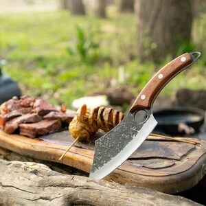 May include: A close-up of a large,  black,  hand-forged cleaver with a wooden handle. The cleaver is sitting on a wooden cutting board with some cooked meat and potato chips.
