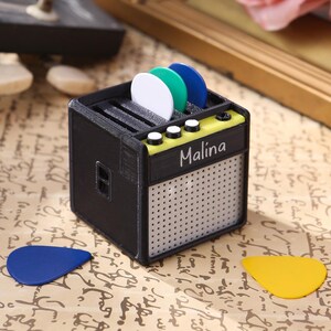 May include: A black and yellow miniature guitar amplifier with the word "Malina" printed on the front. The amplifier has a speaker grille and three guitar picks in a slot on top.